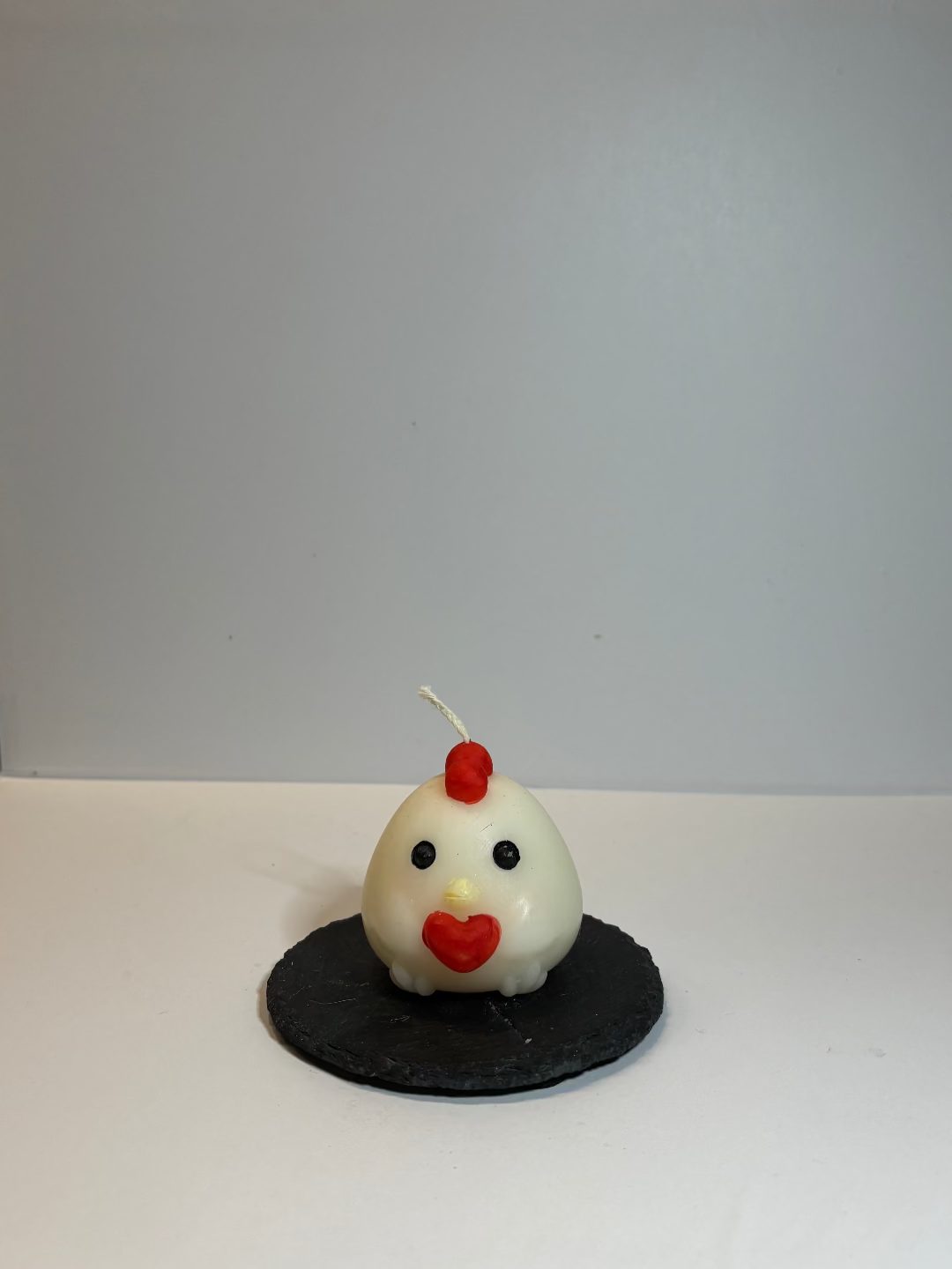 Foto einer Kerze, die aussieht wie ein weißes Küken, mit schwarzen Augen und roter "Mähne", das ein rotes Herz vor sich hält